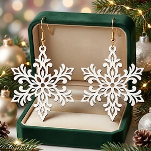 May include: A pair of white snowflake-shaped earrings with intricate cut-out patterns. Each earring features a gold-toned metal hook for attachment. The snowflake design is symmetrical with detailed filigree-like branches extending from the centre.