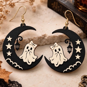 May include: A pair of drop earrings featuring a black crescent moon shape with a cream-coloured ghost wearing a bow, a small hanging lantern, and three stars. The earrings are attached to gold-toned metal hooks for pierced ears.