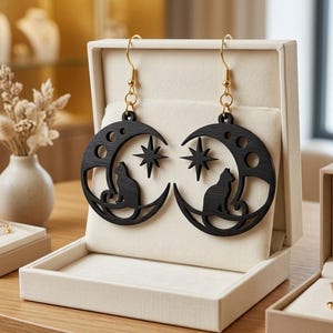 May include: A pair of black wooden drop earrings featuring a crescent moon shape with a cutout cat silhouette and a four-pointed star. The earrings hang from gold-toned metal hooks and are displayed on a cream-colored velvet jewelry stand.