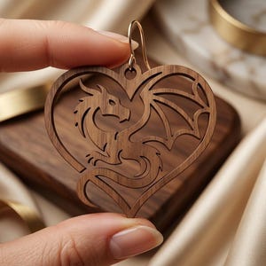 May include: A heart-shaped wooden earring featuring a laser-cut dragon silhouette. The brown wood grain is visible throughout the design, which includes a curved metal hook at the top for suspension. The dragon is depicted in profile with wings.