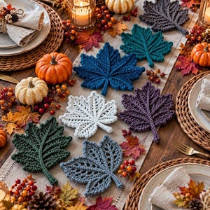 May include: A collection of crocheted leaf-shaped coasters in various colors including dark blue, teal, white, purple, dark green, and grey, arranged on a table runner alongside small pumpkins and decorative autumn foliage.