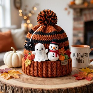 May include: A crocheted beanie with orange and black horizontal stripes, topped with a matching pom-pom. The front features small crocheted appliques of a black spider, a white ghost, a snowman, and colorful autumn leaves.