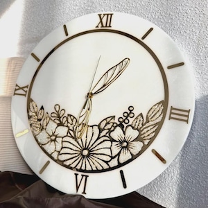 May include: A round white wall clock featuring gold-colored Roman numerals and gold-colored hands shaped like insect wings. The lower portion of the clock face is decorated with a detailed floral design in gold and white textured accents.