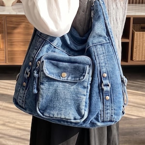 May include: A blue denim hobo bag featuring a washed texture, a single wide shoulder strap, and a front exterior pocket with a metallic snap button closure. The bag includes vertical stitched paneling, side metal hardware accents, and a zipper.