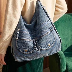 May include: A blue denim hobo bag featuring a slouchy silhouette, multiple zippered compartments, and two front pockets with knotted drawstring closures and metallic snap buttons. The bag includes a single shoulder strap and silver-tone hardware.