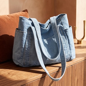 May include: A light blue denim tote bag featuring two long shoulder straps, a spacious main compartment, and visible side pockets. The bag is constructed from a soft, washed-look fabric with reinforced stitching along the seams and handles.