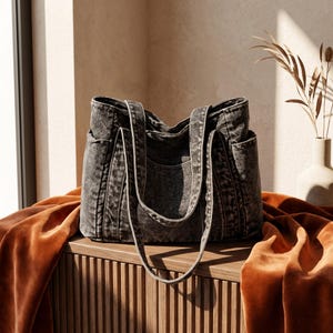 May include: A charcoal gray tote bag with a washed, distressed texture. The bag features two long shoulder straps, a central main compartment, and visible side pockets. The fabric has a mottled, uneven color pattern throughout the exterior.