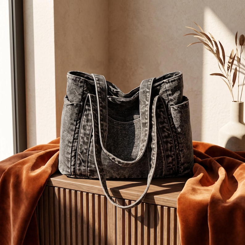 May include: A charcoal gray tote bag with a washed, distressed texture. The bag features two long shoulder straps, a central main compartment, and visible side pockets. The fabric has a mottled, uneven color pattern throughout the exterior.