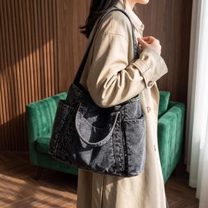 May include: A dark gray acid-wash denim tote bag featuring a spacious main compartment, two sturdy shoulder straps, and two external side pockets. The bag is constructed from a textured fabric with visible stitching details along the seams and edges.