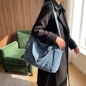 May include: A medium-sized blue denim tote bag featuring two short top handles and a long shoulder strap. The bag has a structured rectangular shape with visible exterior side pockets and reinforced stitching along the seams and handle attachments.