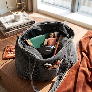 May include: A charcoal gray acid-wash denim tote bag filled with various personal items including a green wallet, a pink perfume bottle labeled Elixir, a black Tom Ford compact, a brown pen, a wristwatch, and a small brown velvet drawstring pouch.