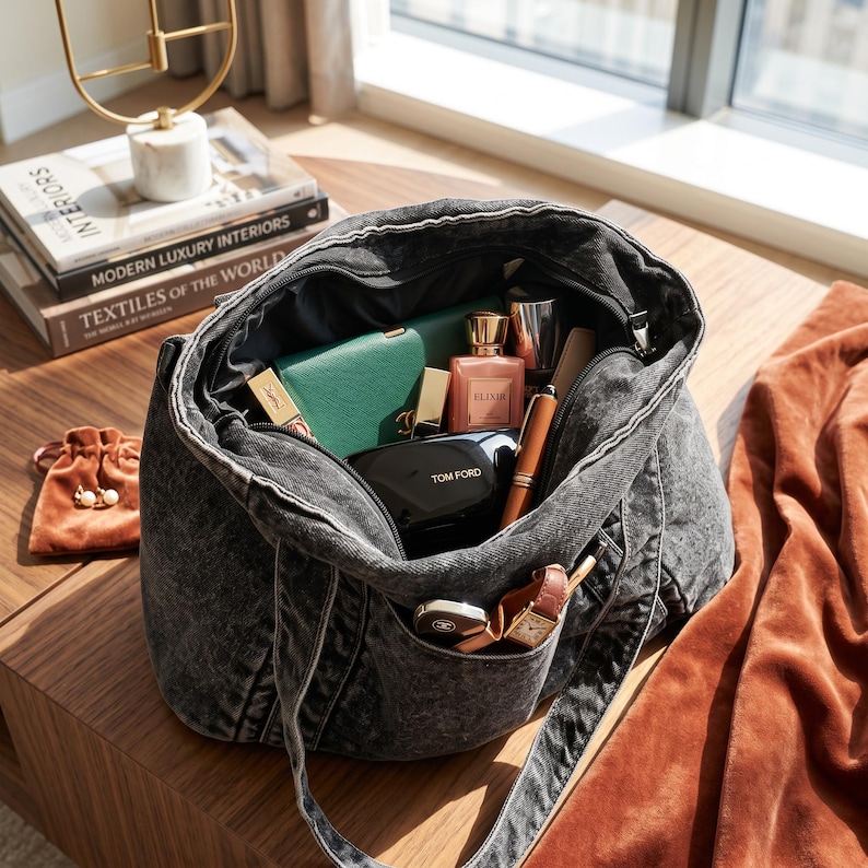 May include: A charcoal gray acid-wash denim tote bag filled with various personal items including a green wallet, a pink perfume bottle labeled Elixir, a black Tom Ford compact, a brown pen, a wristwatch, and a small brown velvet drawstring pouch.