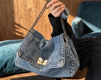 May include: A medium-wash blue denim shoulder bag featuring a fold-over flap with a rectangular gold-tone metal turn-lock closure. The bag includes a metallic chain strap and a matching denim shoulder pad for comfort, showcasing a classic textile design.