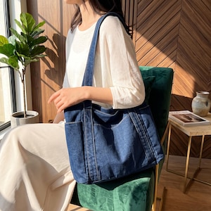 May include: A dark blue denim tote bag featuring a rectangular shape, two long shoulder straps, and two exterior side pockets. The bag is constructed with visible stitching along the seams and edges, providing a structured appearance.
