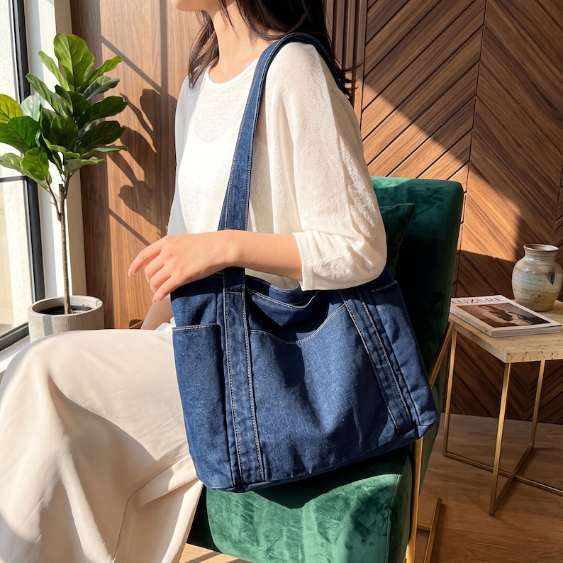 May include: A dark blue denim tote bag featuring a rectangular shape, two long shoulder straps, and two exterior side pockets. The bag is constructed with visible stitching along the seams and edges, providing a structured appearance.