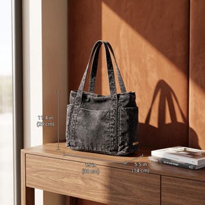 May include: A charcoal gray canvas tote bag with two top handles and exterior side pockets. The bag measures 11.4 inches in height, 15 inches in width, and 5.5 inches in depth. It features a structured rectangular base and a front slip pocket.