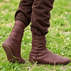 May include: A pair of dark brown leather ankle boots featuring a side-lacing design with thin matching laces. The boots have a smooth finish and are worn with dark brown trousers that have a small cross-stitch detail at the lower hem.