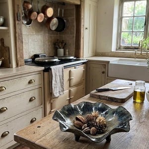 May include: A dark, wavy-edged ceramic bowl with a metallic, iridescent finish sits on a wooden surface. The bowl contains various dried seed pods and pinecones. A small rectangular tag on the table reads HANDMADE ARTISAN POTTERY.