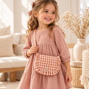 May include: A dusty rose long-sleeved dress with a tiered skirt and a matching hair bow. A small crossbody bag with a white and orange gingham pattern, featuring a ruffled edge and a single silver snap closure, is worn across the chest.