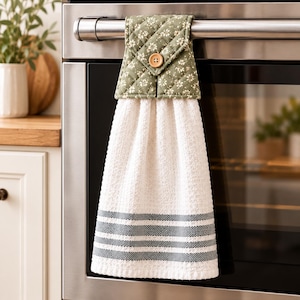 May include: A hanging kitchen towel featuring a white waffle-weave fabric with three dark gray horizontal stripes at the bottom. The top section is a quilted sage green fabric with a small white floral pattern, secured by a single wooden button.