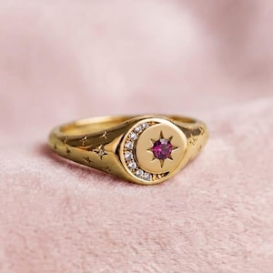 Puede incluir: Un anillo de tono dorado con una cara circular que presenta una piedra preciosa de color rosa oscuro en el centro, rodeada por una forma de media luna con pequeñas piedras claras. La banda tiene grabados múltiples motivos de estrellas.