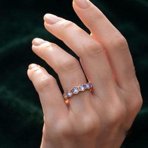 May include: A rose gold band ring featuring a series of connected hexagonal settings, each containing a small, iridescent, light-blue moonstone. The ring has a polished metallic finish and a geometric design that wraps around the finger.