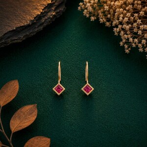 May include: A pair of gold drop earrings featuring a square-cut, vibrant pinkish-red gemstone set in a simple gold frame. The earrings hang from a thin, curved gold wire hook, showcasing a clean, geometric design with a polished metallic finish.