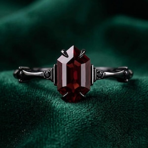 May include: A ring featuring a central elongated hexagonal deep red gemstone set in a dark black metal band. The band is accented with small black stones on either side of the main setting, held by dark prongs that secure the central stone.