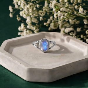 May include: A silver ring featuring a central oval-shaped moonstone with a blue iridescent sheen. The band of the ring splits into two thin, parallel silver wires that meet at the setting of the stone, creating a sleek and open design.