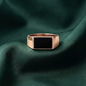 May include: A rose gold signet ring featuring a smooth, rectangular black stone inlay set into the center of the band. The ring has a polished metallic finish and a flat, horizontal orientation, showcasing a clean and simple geometric design.