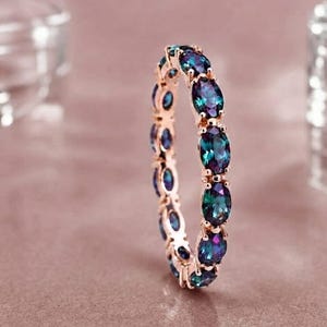 Op de afbeelding: Een eeuwigheidsring van roségoud met een doorlopende rij ovaal geslepen, van kleur veranderende stenen die variëren tussen diepblauwe, groenblauwe en paarse tinten. De gepolijste metalen zetting houdt elke edelsteen vast in een klauwzetting.