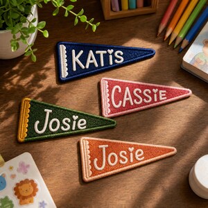Peut inclure: Quatre écussons fanions triangulaires brodés avec des noms cousus en blanc et des cœurs. Les écussons sont bleu marine avec le nom KATIS, rouge avec CASSIE, vert foncé avec Josie et orange avec Josie, chacun avec un bord festonné.