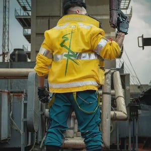 May include: A bright yellow jacket with the word EMERGENCY printed on the collar, a green graphic design on the back, and silver reflective stripes. The jacket is paired with teal trousers featuring yellow suspender straps and a black hand glove.