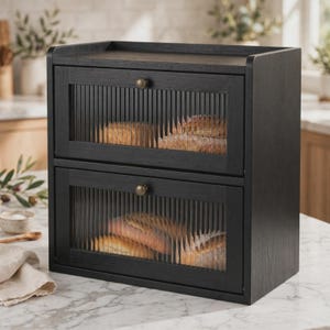May include: A two-tier black wooden bread box featuring two drop-down doors with vertical reeded glass panels and small round brass-toned knobs. The interior compartments contain loaves of bread visible through the textured glass fronts.