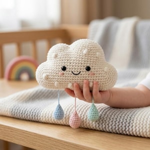 Puede incluir: Un peluche de nube tejido a ganchillo en color crema con una cara sonriente, ojos negros y mejillas rosadas. Tres pequeñas gotas de lluvia tejidas a ganchillo en azul claro, rosa suave y verde menta cuelgan de la nube mediante hilos finos.