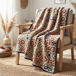 May include: A knitted throw blanket featuring a repeating geometric pattern in shades of rust orange, sage green, cream, and navy blue. The blanket has a dark navy blue border and is draped over a light-colored wooden armchair with a fabric seat.