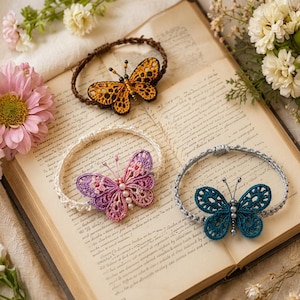 May include: Three bracelets featuring intricate crochet butterfly motifs. One bracelet has a brown band with an orange and black butterfly, one has a white band with a purple and pink butterfly, and one has a silver band with a teal butterfly.
