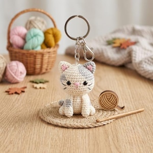 May include: A crochet cat keychain featuring a cream and grey patterned body with small pink ears, a pink nose, and black eyes. The cat is attached to a silver metal split ring and lobster clasp, designed for use as a decorative accessory.