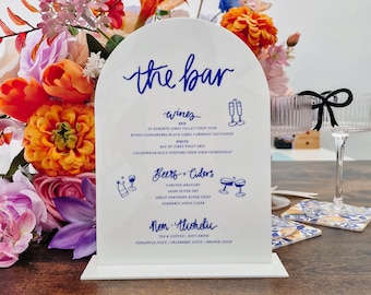May include: A white sign with blue lettering that reads "the bar" and lists various wine, beer, cider, and non-alcoholic beverage options. The sign is on a wooden table with a floral arrangement in the background.