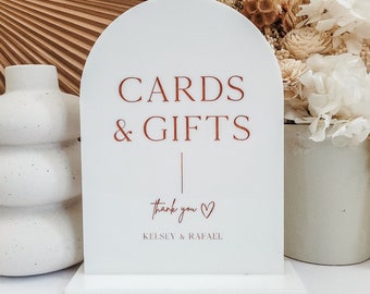 May include: White arch-shaped sign with the text "CARDS & GIFTS" in red. Below the text is a thin black line and the words "thank you" in cursive with a red heart. The text "Kelsey & Rafael" is below the heart in red.