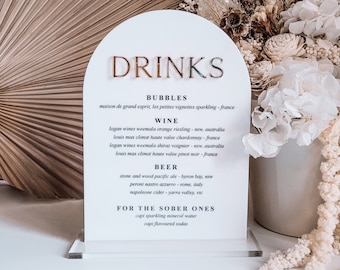 May include: White acrylic sign with a gold arch and the word "DRINKS" in gold lettering. The sign lists different types of drinks, including bubbles, wine, beer, and options for those who prefer non-alcoholic beverages.