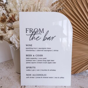 May include: A white arched acrylic sign with black text reading FROM the bar, listing categories for WINE, BEER & CIDER, SPIRITS, and NON ALCOHOLIC with various drink options including sparkling brut, sauvignon blanc, and others.