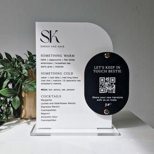 May include: A white arched acrylic menu sign with black text listing drink options for Sarah Kae Hair. A black oval plaque is attached to the right side with gold hardware, featuring a QR code and the text Let's Keep In Touch Bestie.