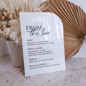 May include: A white arched acrylic sign with black text reading FROM the bar, listing categories for wine, beer and cider, spirits, and non alcoholic drinks. The sign features a smooth, glossy finish and a clean, modern, semi-circular top edge.