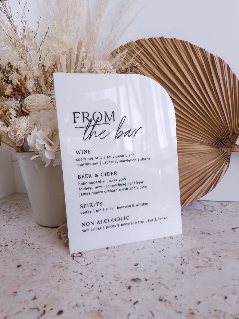 May include: A white arched acrylic sign with black text reading FROM the bar, listing categories for wine, beer and cider, spirits, and non alcoholic drinks. The sign features a smooth, glossy finish and a clean, modern, semi-circular top edge.