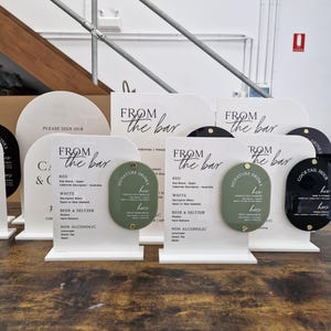 May include: White acrylic arched signs with black text reading FROM THE BAR and lists of drink options. Each sign features an oval-shaped sage green or black accent piece attached with gold hardware, labeled SIGNATURE DRINKS with his and hers options.