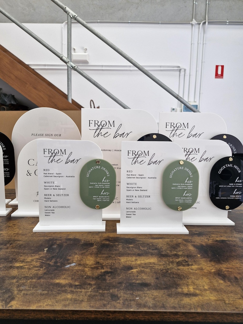May include: White acrylic arched signs with black text reading FROM THE BAR and lists of drink options. Each sign features an oval-shaped sage green or black accent piece attached with gold hardware, labeled SIGNATURE DRINKS with his and hers options.
