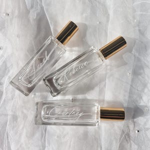 May include: Three clear glass bottles with gold caps and personalized names: Sarah, Rebecca, and Courtney. The bottles are on a white fabric background.