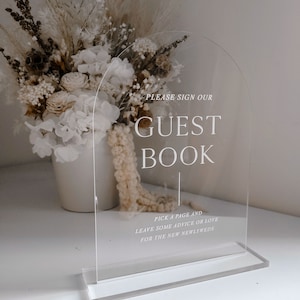 May include: Transparent acrylic guest book sign with an arched top and rectangular base. The sign reads "PLEASE SIGN OUR GUEST BOOK" with additional text below. A bouquet of white flowers is in the background.