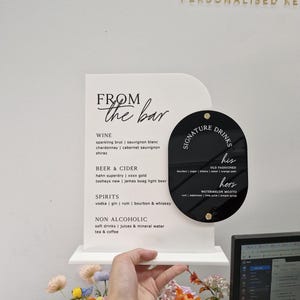 May include: A white arched acrylic sign with black text titled FROM the bar, listing wine, beer, cider, spirits, and non-alcoholic options. Attached is a black oval acrylic sign titled SIGNATURE DRINKS with his and hers cocktail recipes.
